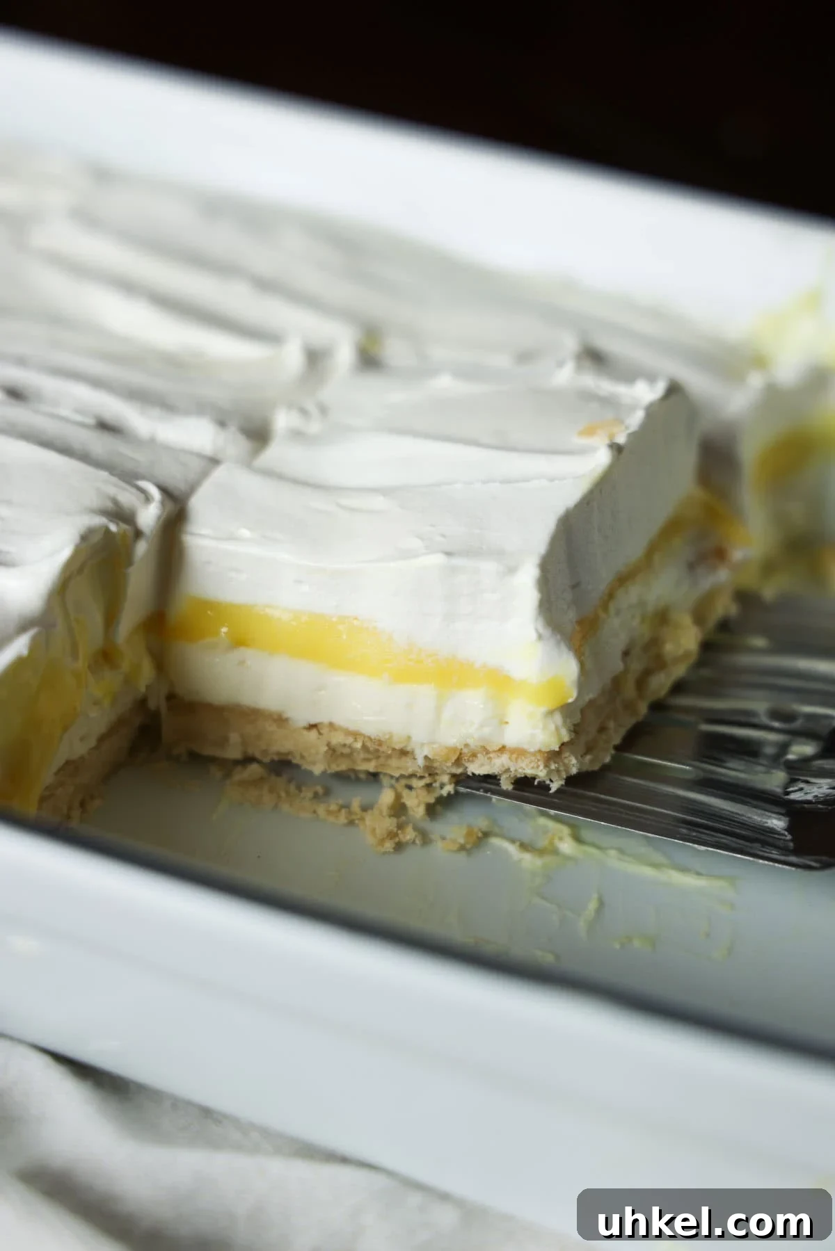 These super easy, nostalgic Lemon Lush is the perfect make-ahead dessert. Layers of creamy whipped topping, lemon pudding, and cheesecake all topped on the most delicious, buttery shortbread crust! A spatula serving a slice of layered Lemon Lush dessert in a white ceramic dish.