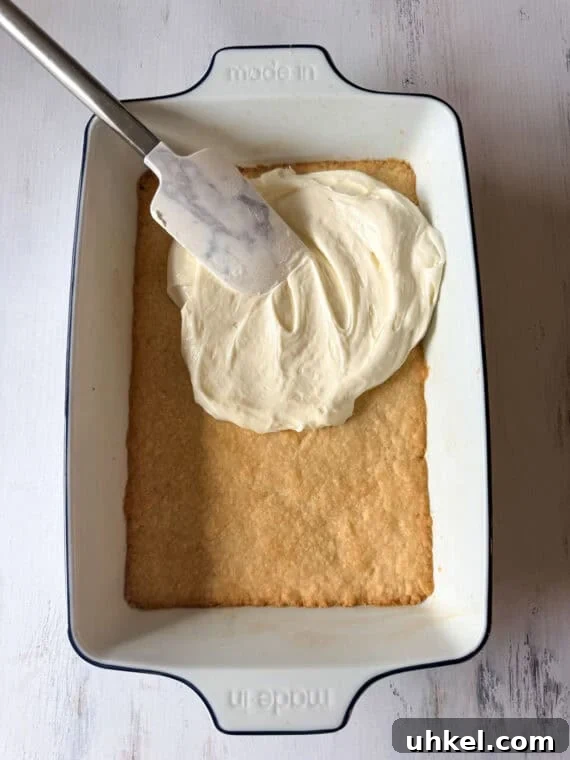 Decadent Lemon Lush Dessert with Graham Cracker Crust 3 A shortbread crust in a white baking pan being topped with a cream cheese layer for a Lemon Delight dessert.
