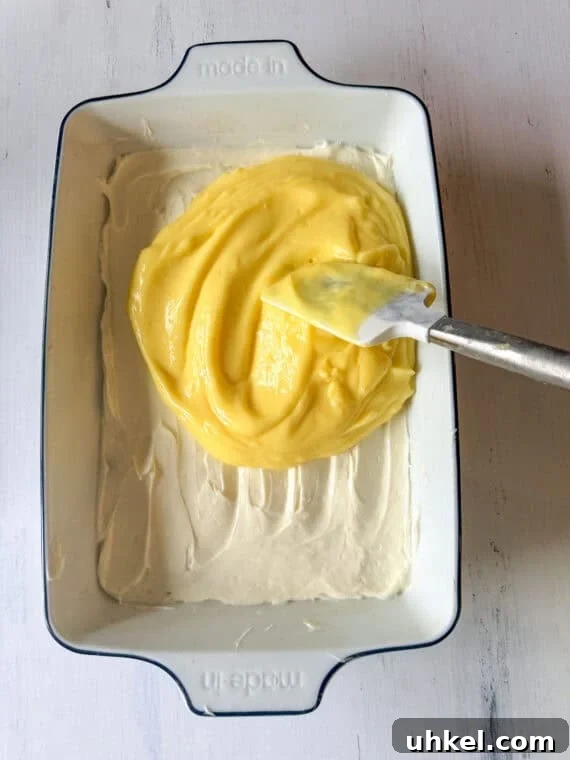 Decadent Lemon Lush Dessert with Graham Cracker Crust 5 Assembling Lemon Lush dessert by spreading lemon pudding on top of a cream cheese layer in a ceramic pan.