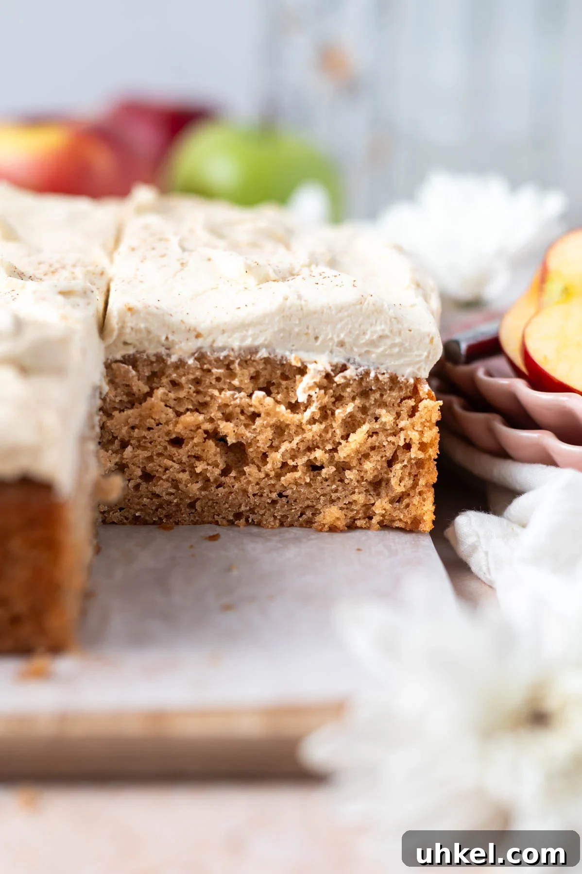 Apple Cider Snack Cake with Tender Apples 3 slice of cake with frosting on a parchment lined cutting board with red and green apples in the background