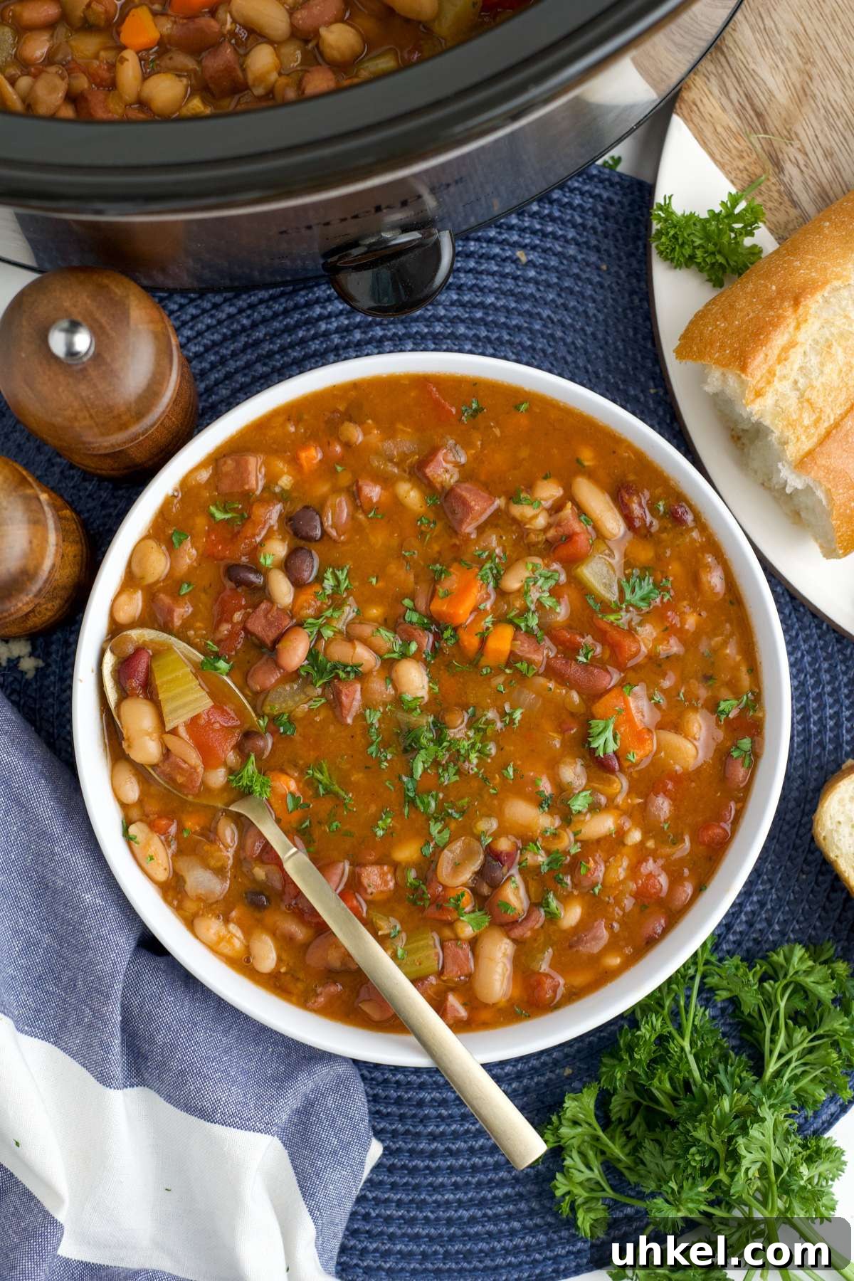 Hearty Slow Cooker 15-Bean Soup Recipe for Meal Prep 2 A bowl of slow cooker 15 bean soup on the table with a spoon in the bowl and a bowl of bread and salt and pepper shakers to the side.