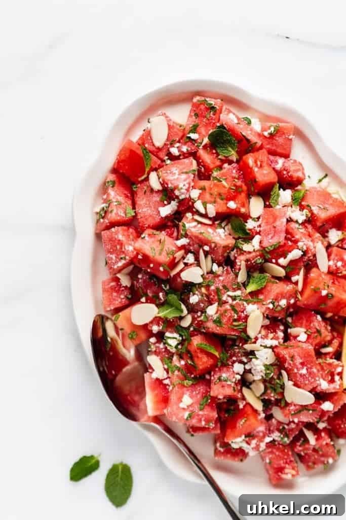 cubed watermelon with slivered almonds, feta cheese, and chopped mint leaves on a white serving platter on a white marble countertop, summer salads with watermelon