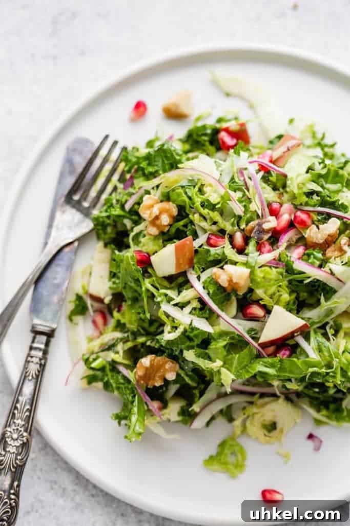 summer salads made with shaved brussels sprouts, kale, fennel, and chopped apples topped with walnuts and pomegranate seeds on a glossy white plate with a silver fork and knife