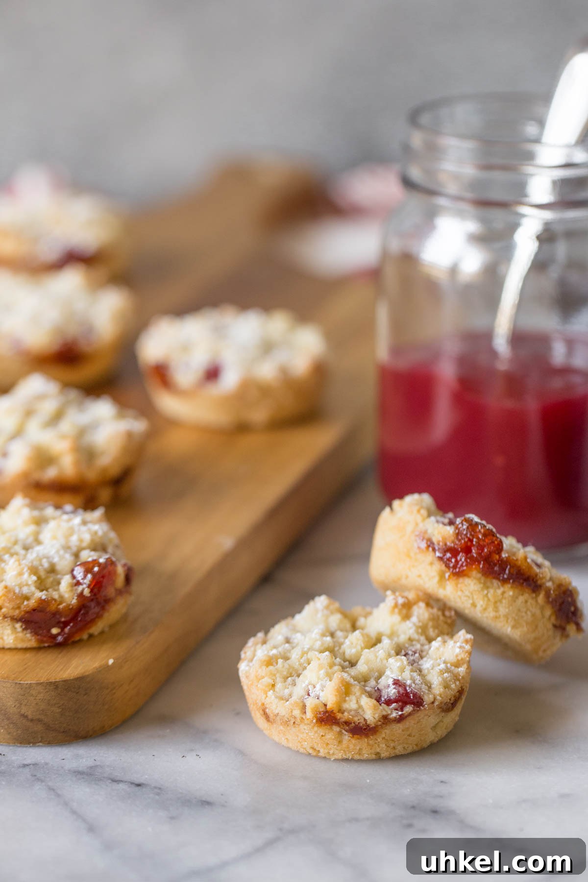 Costco Raspberry Crumble Cookies Review & Where to Buy 4 Two Costco Raspberry Crumble Cookies with a jar of raspberry preserves in the background, along with more cookies on a wooden cutting board.