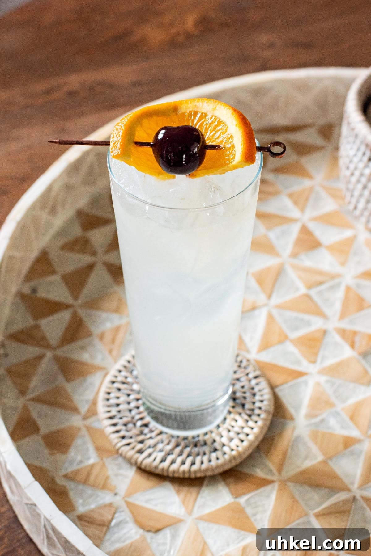 Classic Tom Collins Cocktail Recipe: How to Make a Refreshing Gin Collins 2 A Tom Collins cocktail garnished with an orange slice and maraschino cherry, pictured on a wooden tray and table.