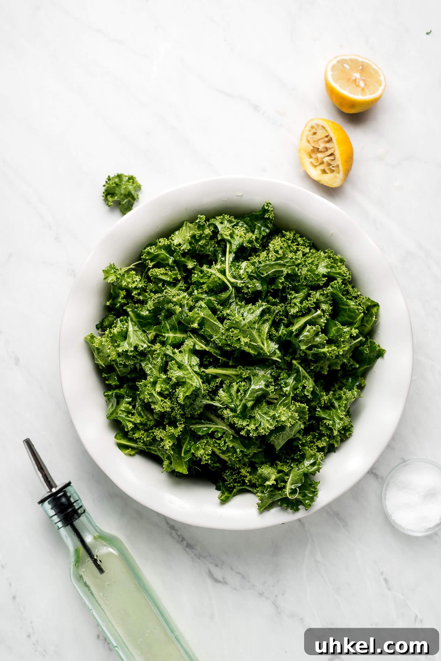 How to Massage Kale for Tender, Flavorful Salads 2 Massaged Kale in a shallow bowl with a squeeze lemon, bottle of oil, and bowl of salt to the sides.