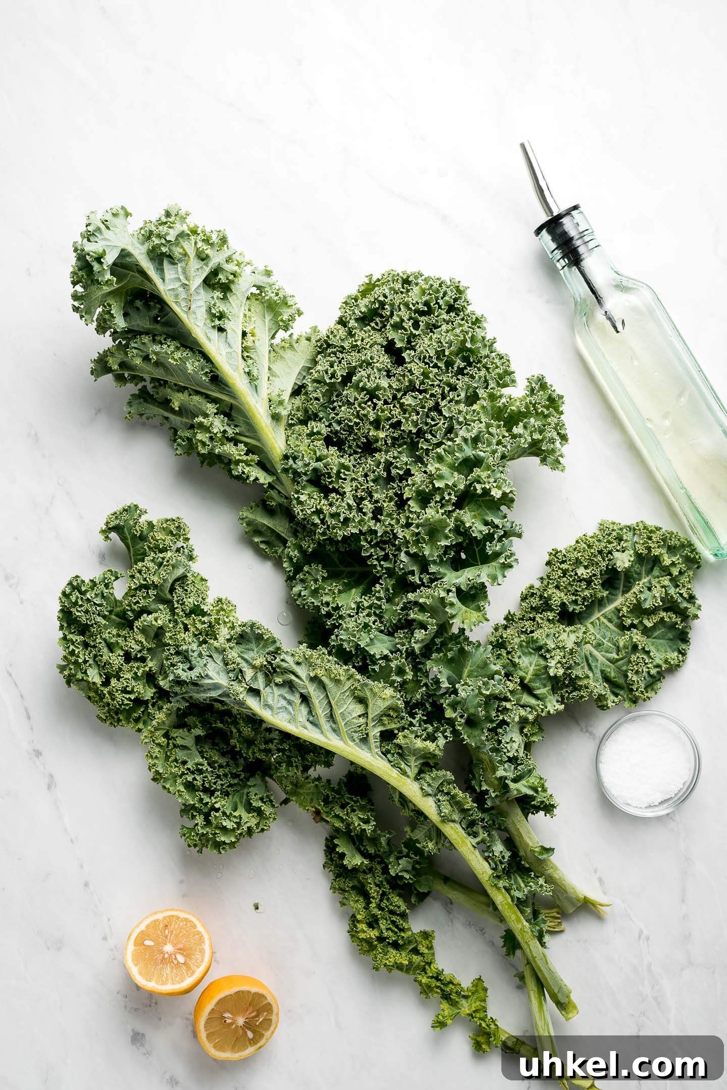 How to Massage Kale for Tender, Flavorful Salads 5 Leaves of raw kale on a marble surface with a bottle of oil, lemon halves, and a small bowl of kosher salt.