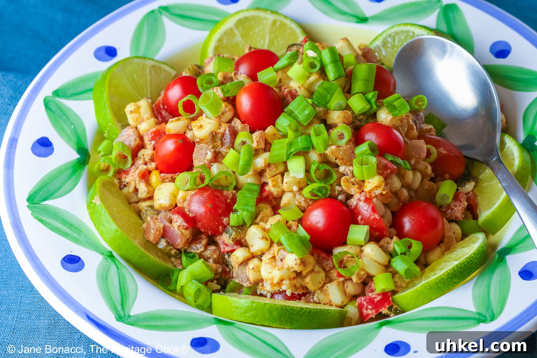 Freshly cut corn tossed with sauteed vegetables and spicy seasoned creamy sauce with red cherry tomatoes and green onions on top, lime slices around the edges of the bowl © 2024 Jane Bonacci, The Heritage Cook.