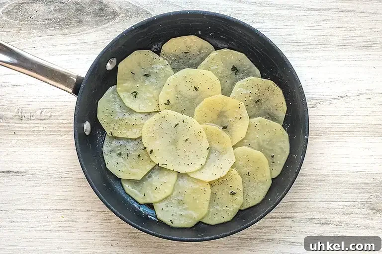 Classic Pommes Anna: Crispy French Layered Potato Cake 5 Thinly sliced potatoes cooking in a skillet with herbs and black pepper.