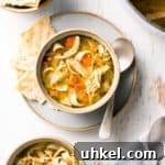 A bowl of gluten free chicken noodle soup shown from above with crackers and a spoon to the side.