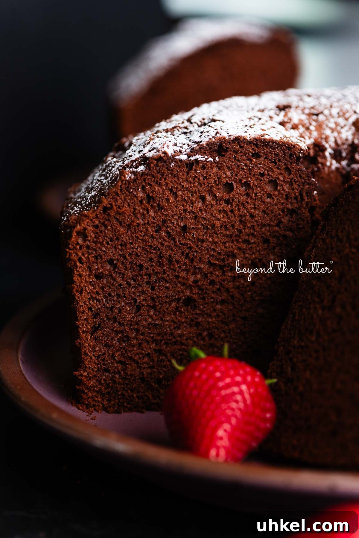 Moist Chocolate Chiffon Cake Recipe for Light, Fluffy Dessert 2 Chocolate chiffon cake sliced with whole strawberries placed on and in front of a dark purple ceramic plate on a black background.