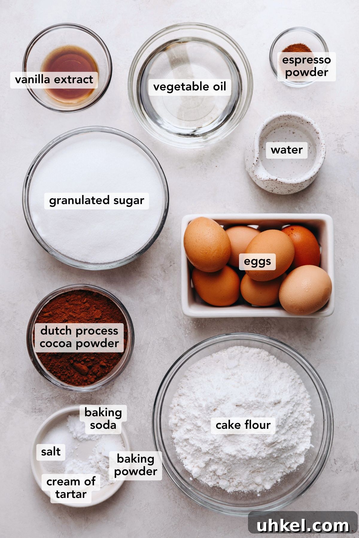 Moist Chocolate Chiffon Cake Recipe for Light, Fluffy Dessert 4 Ingredients in various size plates and bowls labeled as vanilla extract, vegetable oil, espresso powder, granulated sugar, water, eggs, dutch process cocoa powder, cake flour, baking soda, baking powder, cream of tartar, and salt on a white marbled background.