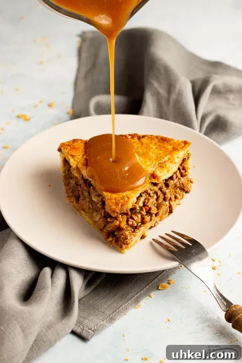 A slice of vegetarian mince and onion pie on a light grey plate with a wooden handled fork resting on it and gravy being poured onto it. Set on a grey linen napkin over a pale blue wash backdrop.