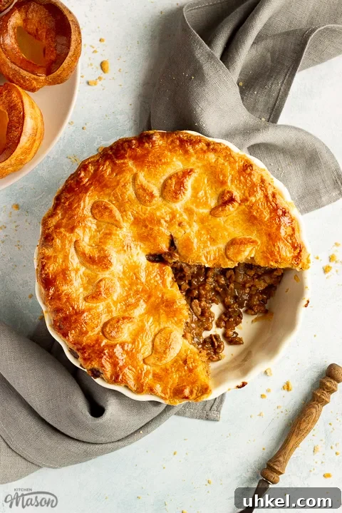 A baked vegetarian mince and onion pie with a slice cut out, decorated with pastry leaves, sat on a grey linen napkin. There's a wooden handled fork and a plate of Yorkshire puddings in the background. Set on a pale blue wash backdrop.