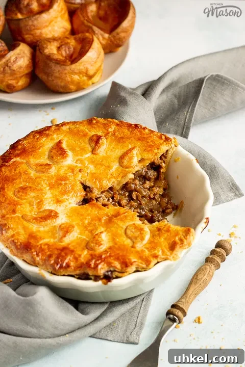 A baked vegetarian mince and onion pie with a slice cut out, decorated with pastry leaves, sat on a grey linen napkin. There's a wooden handled fork and a plate of Yorkshire puddings in the background. Set on a pale blue wash backdrop.