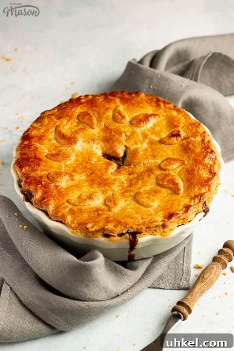 A baked vegetarian mince and onion pie decorated with pastry leaves sat on a grey linen napkin with a wooden handled fork. Set on a pale blue wash backdrop.