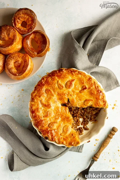 A baked vegetarian mince and onion pie with a slice cut out, decorated with pastry leaves, sat on a grey linen napkin. There's a wooden handled fork and a plate of Yorkshire puddings in the background. Set on a pale blue wash backdrop.