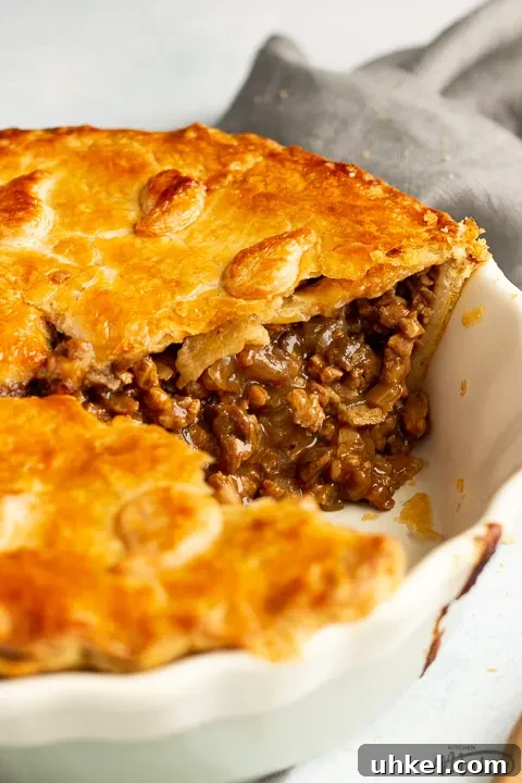A baked vegetarian mince and onion pie with a slice cut out, decorated with pastry leaves, sat on a grey linen napkin. Set on a pale blue wash backdrop.