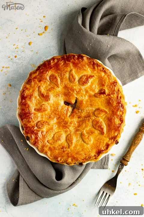 A baked vegetarian mince and onion pie decorated with pastry leaves sat on a grey linen napkin with a wooden handled fork. Set on a pale blue wash backdrop.