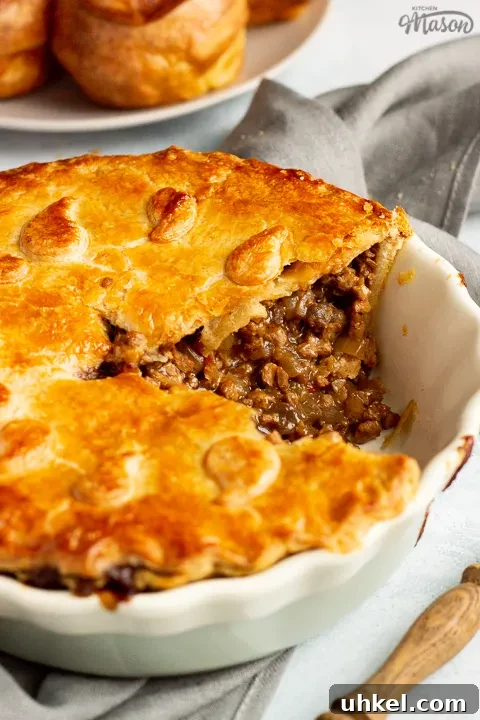 A baked vegetarian mince and onion pie with a slice cut out, decorated with pastry leaves, sat on a grey linen napkin. There's a wooden handled fork and a plate of Yorkshire puddings in the background. Set on a pale blue wash backdrop.
