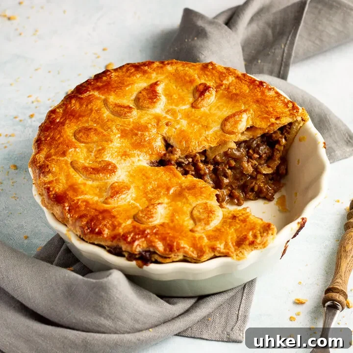 A baked vegetarian mince and onion pie with a slice cut out, decorated with pastry leaves, sat on a grey linen napkin with a wooden handled fork. Set on a pale blue wash backdrop.