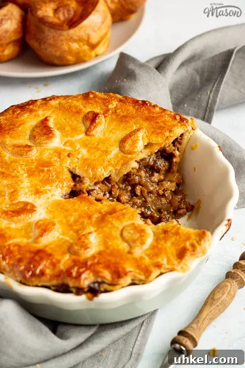 A baked vegetarian mince and onion pie with a slice cut out, decorated with pastry leaves, sat on a grey linen napkin. There's a wooden handled fork and a plate of Yorkshire puddings in the background. Set on a pale blue wash backdrop.