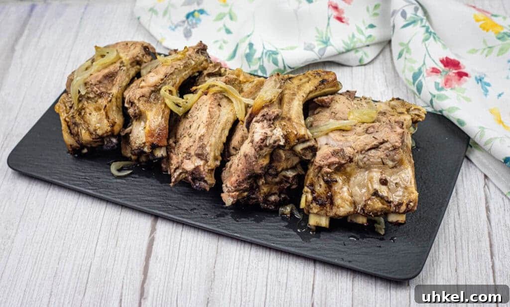 35 Timeless Southern Recipes That Always Delight 32 Oven Baked Ribs with cooked onions on a black rectangular slate platter, set on a wooden surface. A floral-patterned cloth is partially visible in the background.