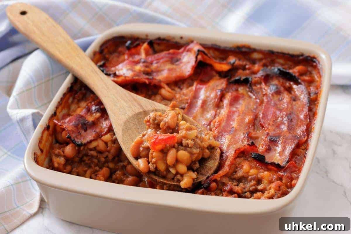35 Timeless Southern Recipes That Always Delight 35 A square baking dish filled with Southern Ground Beef & Baked Bean Casserole and topped with strips of cooked bacon; a wooden spoon holds a serving.