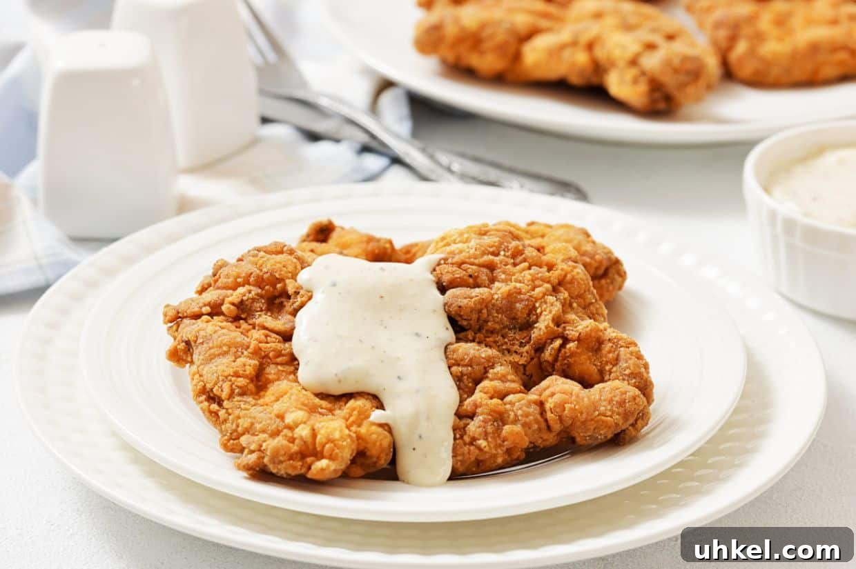 35 Timeless Southern Recipes That Always Delight 7 A plate of fried chicken steak topped with white gravy, with more fried pieces and a bowl of gravy in the background.