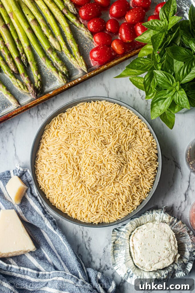 Creamy Orzo with Oven-Roasted Asparagus and Parmesan 3 raw orzo, raw asparagus and tomatoes in a pan, parmesan cheese on blue tea towel, basil leaves on marble board, boursin cheese unwrapped on countertop.