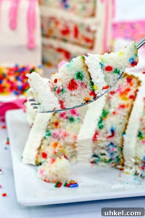 a fork containing a bite of vanilla cake with sprinkles, with the whole cake in the background.
