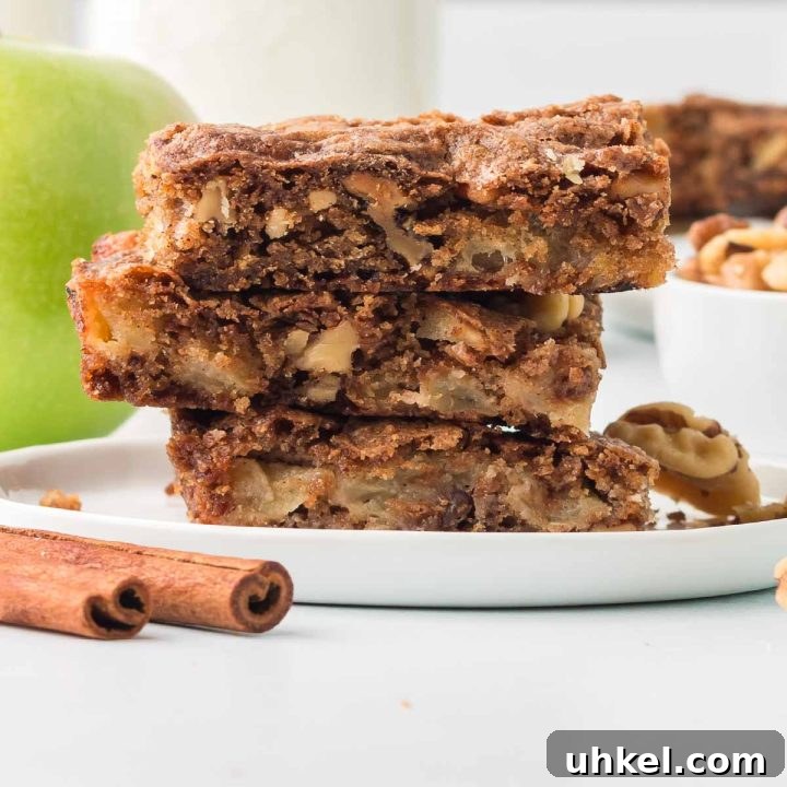 Three Apple Walnut Bars stacked on a white plate with a bowl of walnuts and a green apple in the background.