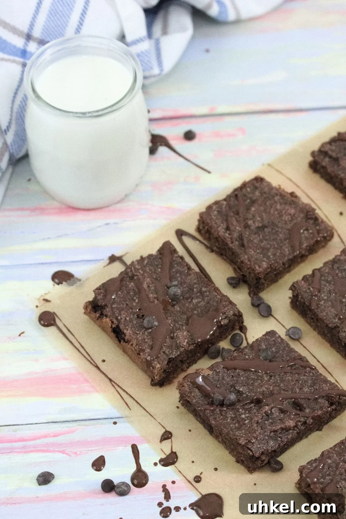 High-Protein Gluten-Free Brownies for Chocolate Lovers 2 Dairy-free chocolate brownies drizzled with chocolate sauce on brown parchment paper with chocolate chips around them and a glass of milk with a blue linen to the side.