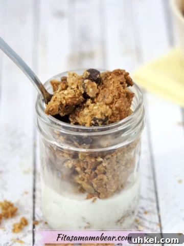 Homemade granola in a small mason jar with milk in it and a spoon in the top. More granola scattered around the jar and in a beige bowl behind it