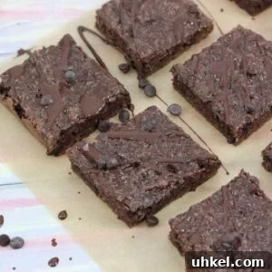 High-Protein Gluten-Free Brownies for Chocolate Lovers 13 Dairy-free chocolate brownies drizzled with chocolate sauce on brown parchment paper with chocolate chips around them and a glass of milk with a blue linen to the side