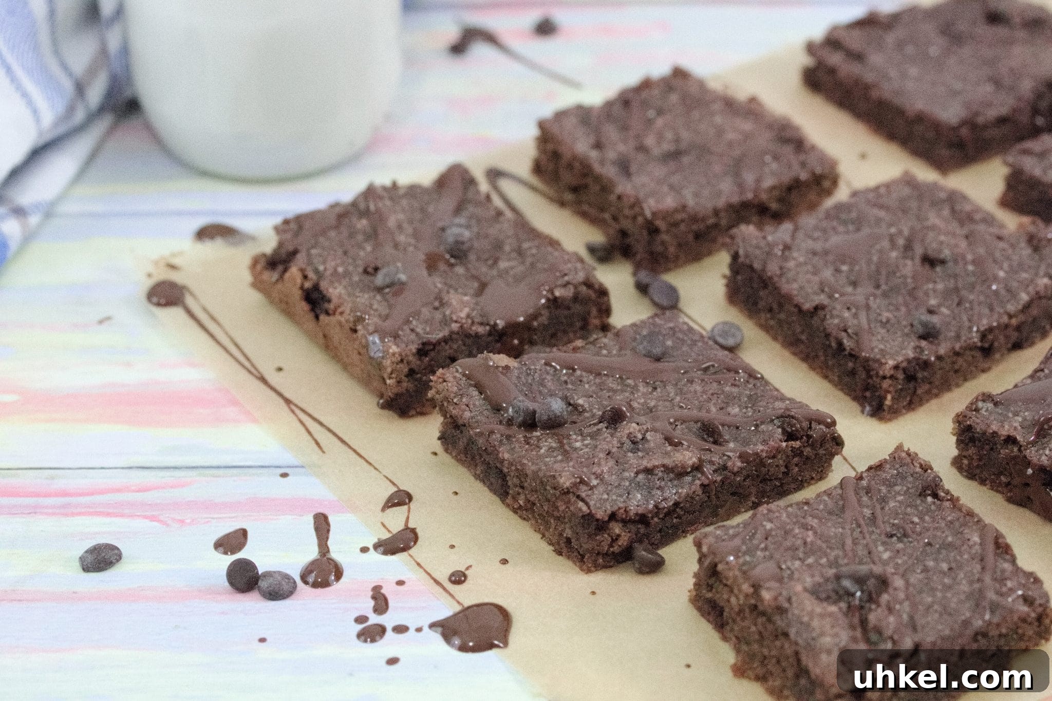 High-Protein Gluten-Free Brownies for Chocolate Lovers 14 Dairy-free chocolate brownies drizzled with chocolate sauce on brown parchment paper with chocolate chips around them and a glass of milk with a blue linen to the side