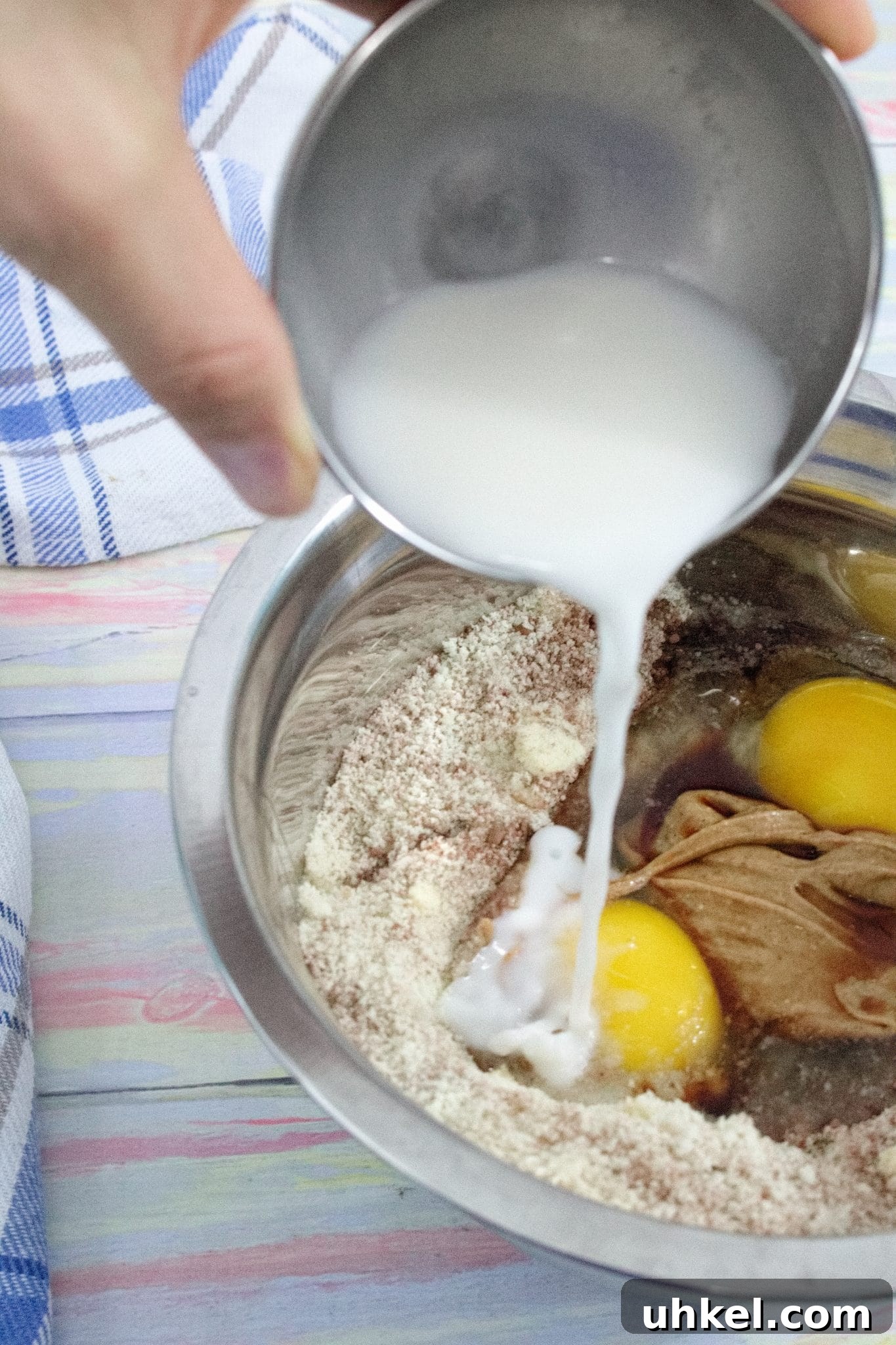 High-Protein Gluten-Free Brownies for Chocolate Lovers 5 Steel bowl with flours, raw eggs and peanut butter in it with a hand pouring in dairy-free milk