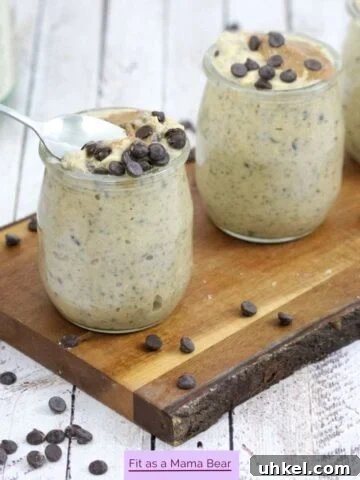 Three mason jars filled with high protein cookie dough overnight oats topped with chocolate chips on a wooden cutting board and a spoon going into one jar.