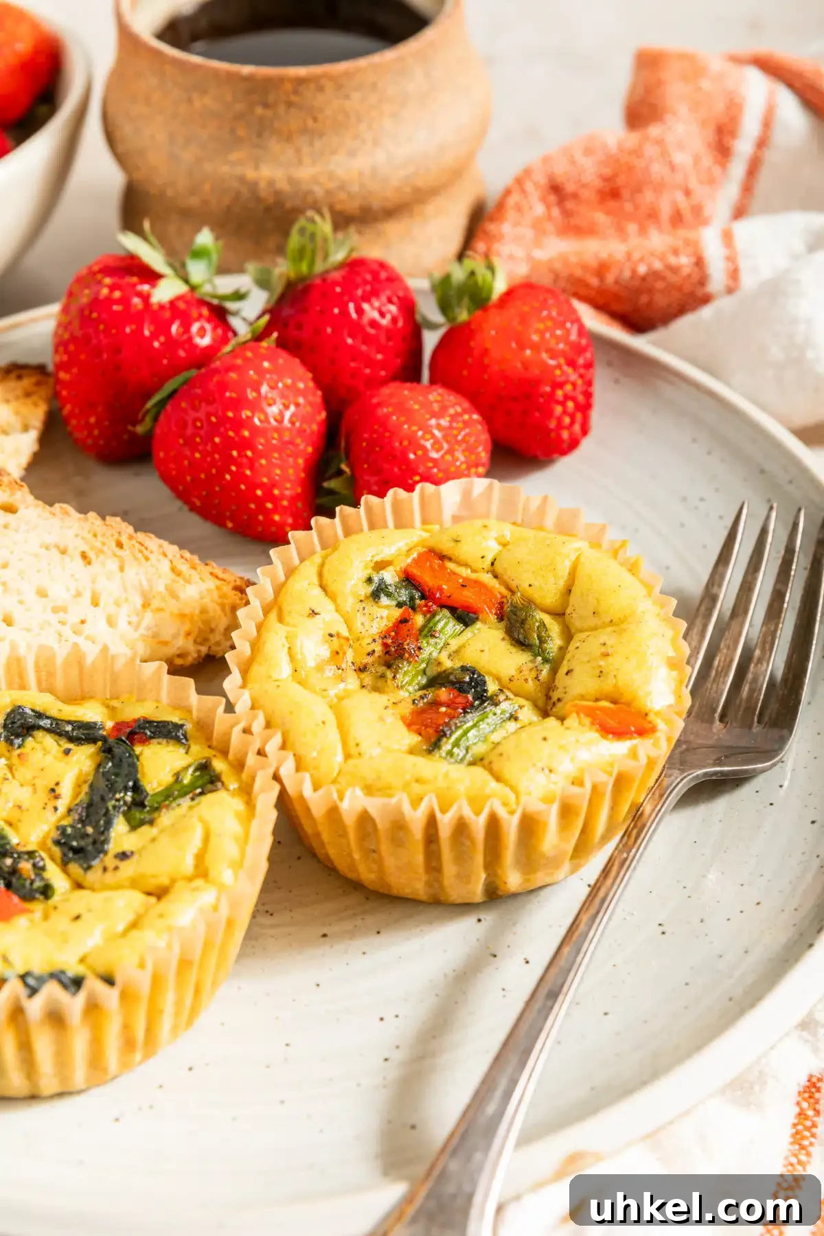Vegan Egg Bites @spabettie #vegan #glutenfree #eggfree #breakfast #recipe Two mini vegan quiche baked in cupcake liners are on a white plate with a fork, strawberries, and toast. The vegan egg bites are bright yellow from turmeric, and have red peppers, spinach, and asparagus in them.