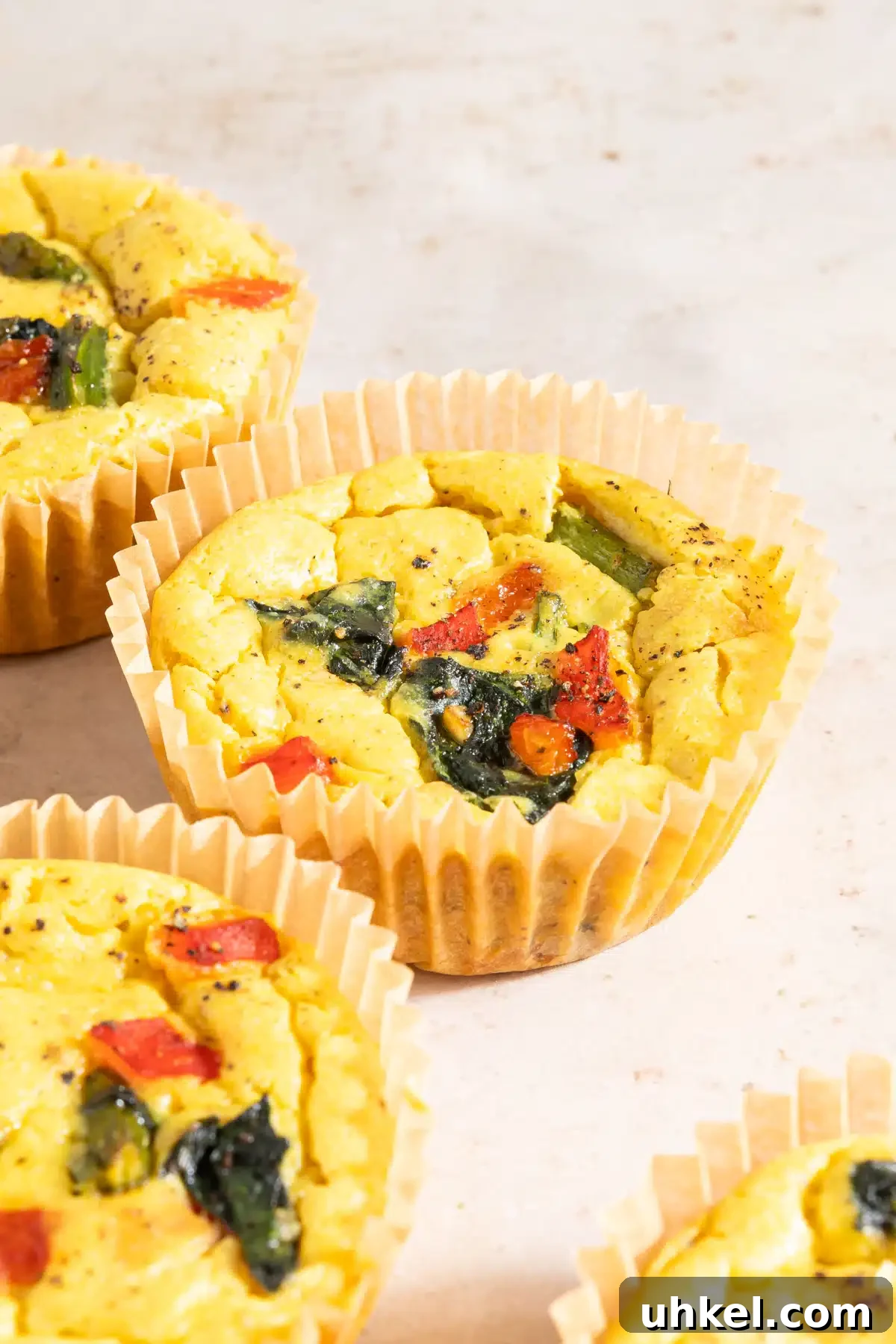 Vegan Egg Bites @spabettie #vegan #glutenfree #eggfree #breakfast #recipe Four mini vegan egg cups baked in cupcake liners sit on an off white surface. The vegan egg bites are bright yellow from turmeric, and are filled with red peppers, spinach, and asparagus.