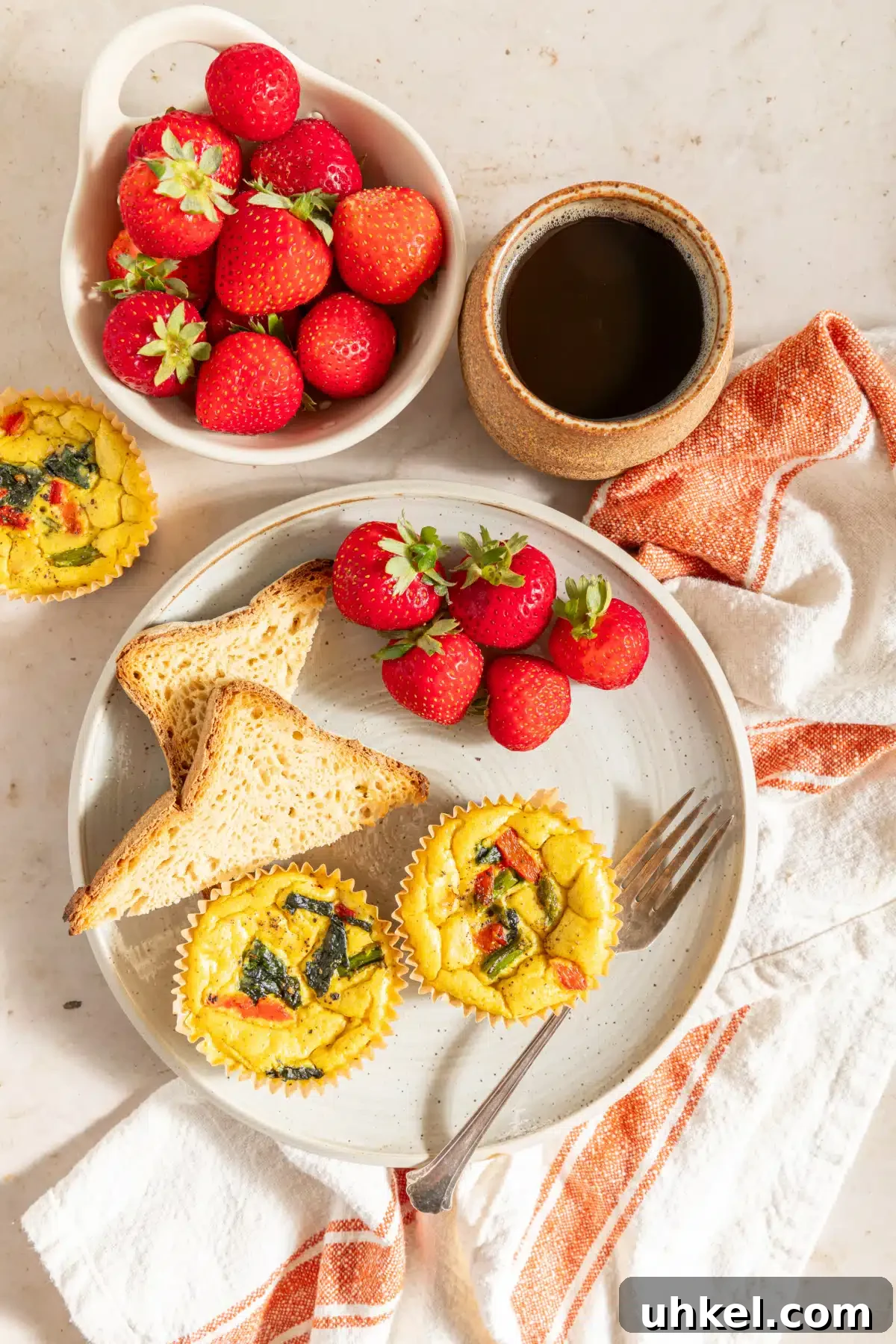 Vegan Egg Bites @spabettie #vegan #glutenfree #eggfree #breakfast #recipe Overhead view of two mini vegan quiche baked in cupcake liners on a white plate with a fork, strawberries, and toast. The vegan egg bites are bright yellow from turmeric, and have red peppers, spinach, and asparagus in them. A bowl of strawberries and a cup of coffee sit next to the plate with a white and orange cloth napkin.