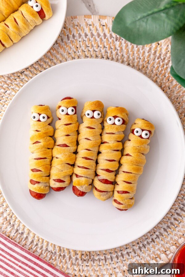 Classic Beanie Weenies Recipe with Flavorful Tomato Sauce 12 Mummy hot dogs on a white plate, with candy eyes.