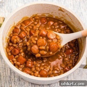 Classic Beanie Weenies Recipe with Flavorful Tomato Sauce 13 Close up photo of beans and weenies in a saucepan with a large spoon in the center.