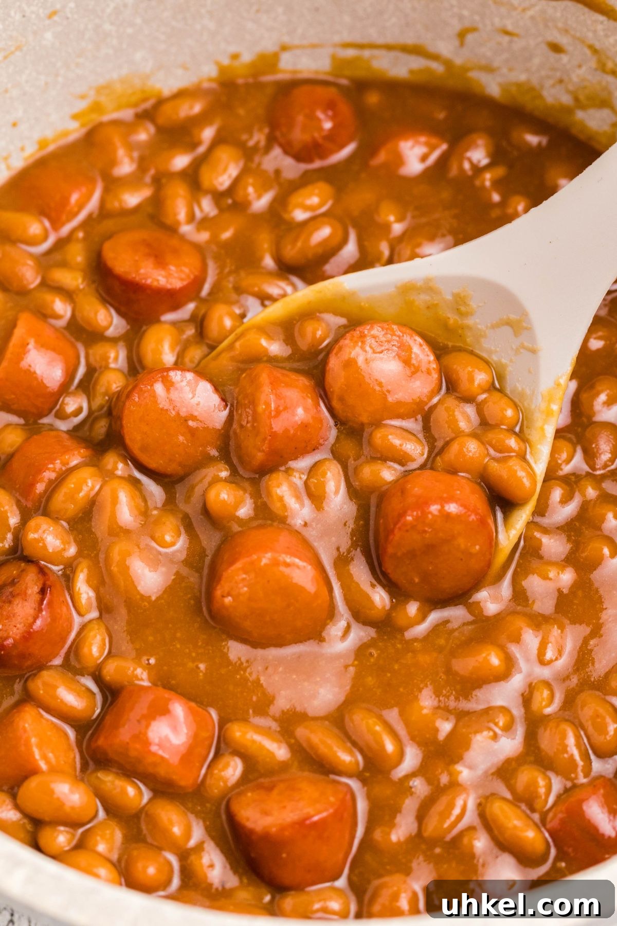 Classic Beanie Weenies Recipe with Flavorful Tomato Sauce 7 Close up photo of beans and weenies in a saucepan with a large spoon in the center.