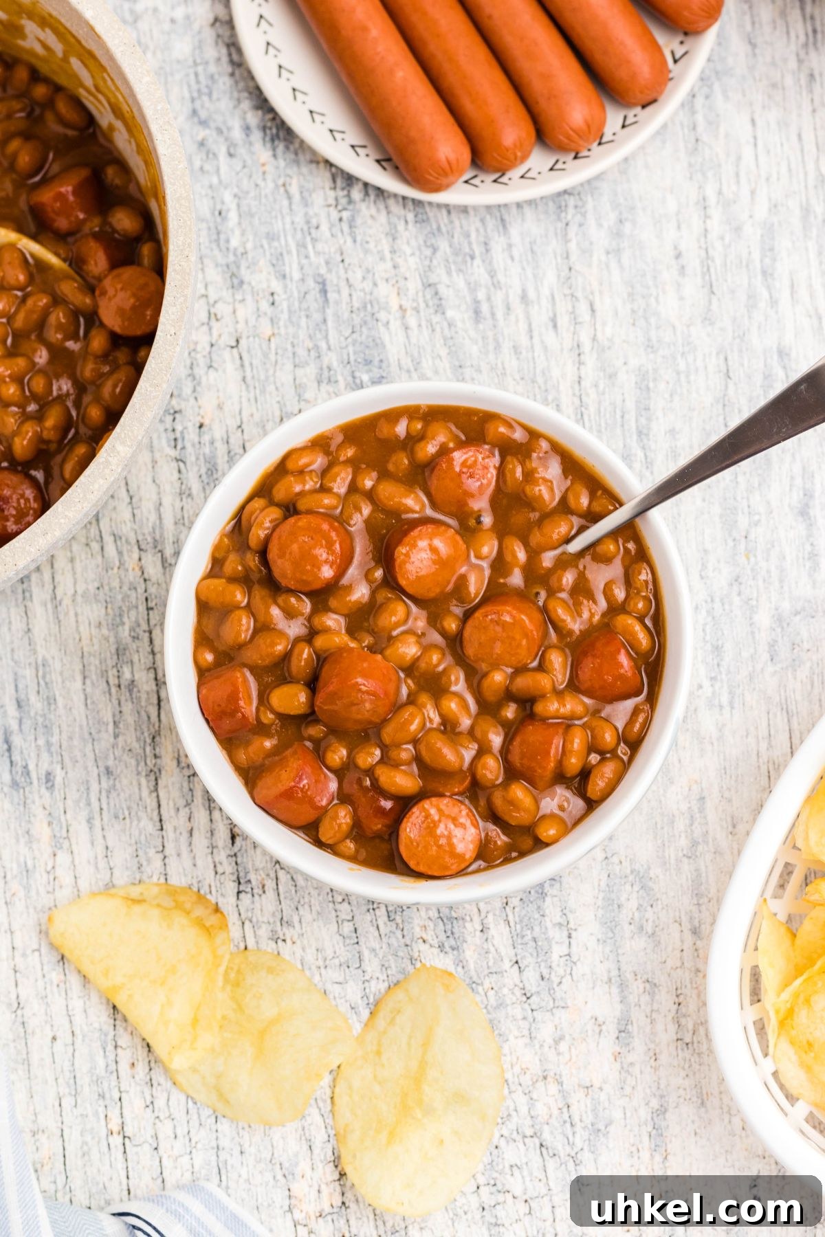 Classic Beanie Weenies Recipe with Flavorful Tomato Sauce 8 Cut up hot dogs, mixed with beans, in a white bowl with potato chips scattered around the dish.