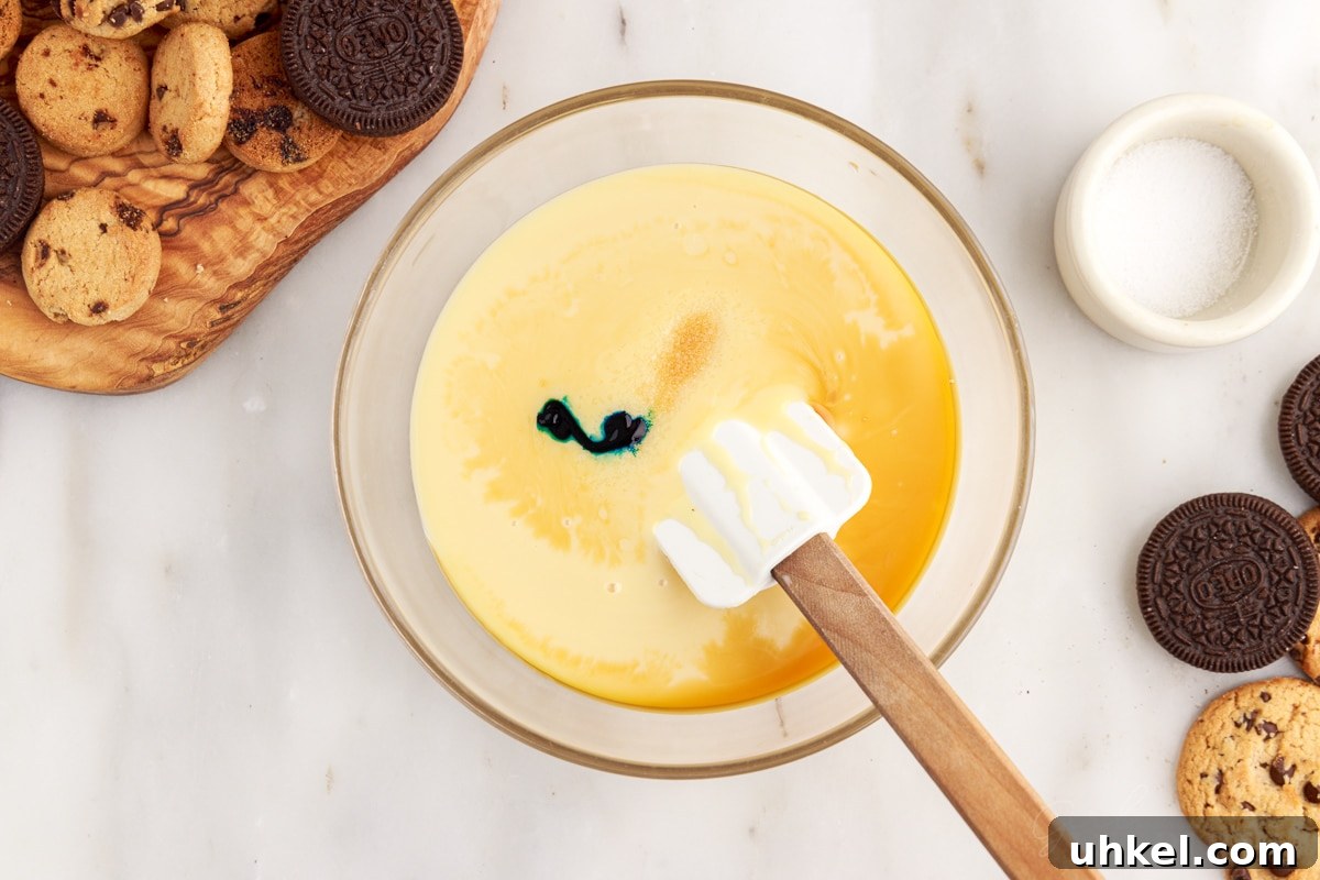 Sweetened condensed milk, salt, vanilla, and blue food coloring in a mixing bowl with a spatula.