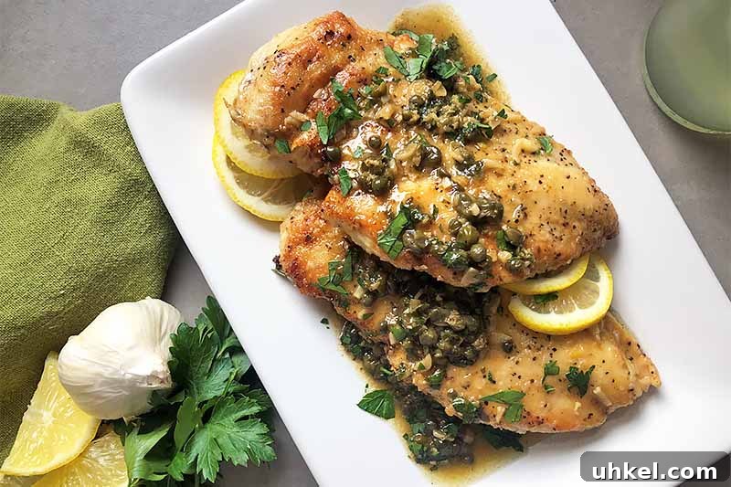Classic Chicken Piccata with Lemon Butter Sauce Recipe 11 Horizontal image of a white plate with cooked poultry pieces covered in a citrus and caper sauce next to whole garlic and lemon slices.