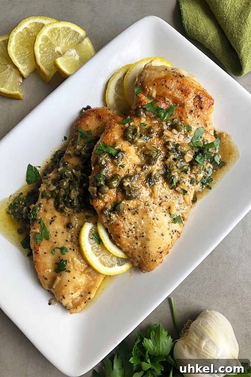 Classic Chicken Piccata with Lemon Butter Sauce Recipe 3 Vertical image of a white plate with cooked poultry pieces covered in a citrus and caper sauce next to whole garlic and lemon wedges.