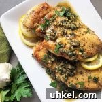 Horizontal image of a white plate with cooked poultry pieces covered in a citrus and caper sauce next to whole garlic and lemon slices.