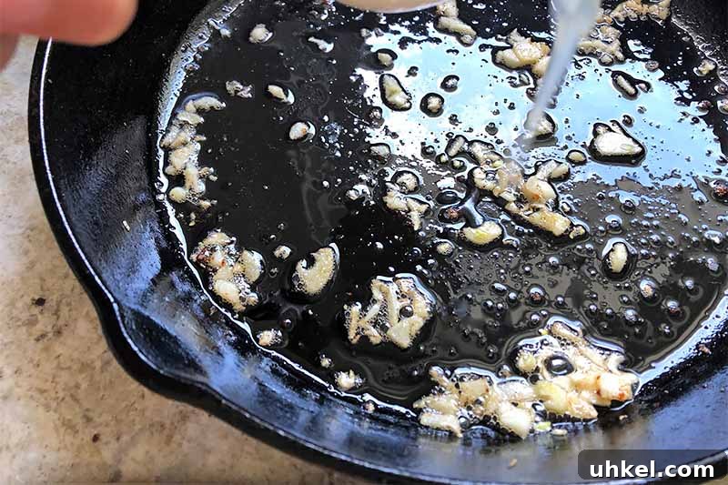 Classic Chicken Piccata with Lemon Butter Sauce Recipe 7 Horizontal image of cooking minced garlic in a cast iron skillet while deglazing with broth.